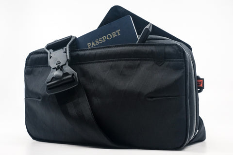 An Alpha Pack shown from the front with passport, pen and phone sticking out and strap with fidlock buckle draped over the front | Color: Black VX | Size: 2.6L